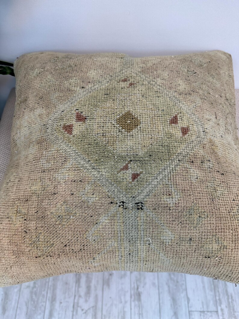 May include: A square throw pillow featuring a textured, woven surface in shades of beige, tan, and light green. The center displays a faded, geometric diamond pattern with small, muted red accents and a central brown dot, creating a worn appearance.