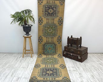 May include: A long, rectangular rug with a beige background and geometric patterns in navy blue, teal, and olive green. The rug features a repeating square design and floral accents, suitable for a hallway or entryway.