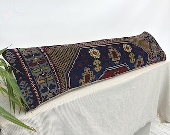 Puede incluir: Un cojín cilíndrico largo y rectangular con una funda de tejido con un campo central azul oscuro, patrones geométricos intrincados en rojo, dorado y gris, y un borde decorativo con motivos repetitivos en un diseño inspirado en alfombras.