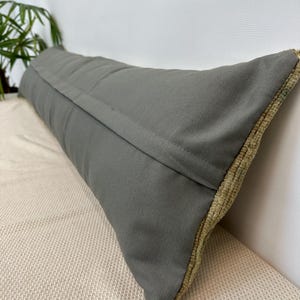 May include: A long, rectangular lumbar cushion featuring a solid dark grey fabric on the front side with a visible central seam. The side edges of the cushion display a textured, light beige woven fabric with a distinct, raised pile construction.