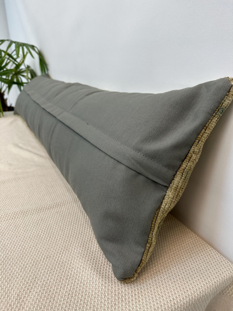 May include: A long, rectangular lumbar cushion featuring a solid dark grey fabric on the front side with a visible central seam. The side edges of the cushion display a textured, light beige woven fabric with a distinct, raised pile construction.