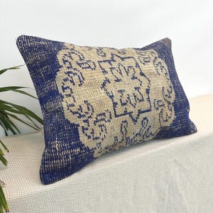 May include: Rectangular decorative pillow with a blue border and a beige center featuring a geometric pattern. The pillow has a textured appearance, suggesting a woven or embroidered design. The color palette is a combination of blue and beige.