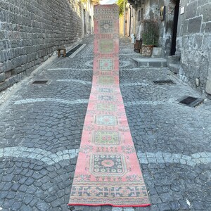 May include: A long, rectangular pink rug with a repeating geometric pattern in green, blue, and cream. The rug is displayed outdoors on a cobblestone street, showcasing its length and design.
