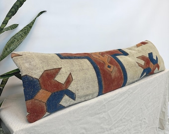 Puede incluir: Un cojín lumbar largo y rectangular con una funda de tejido textil sobre una base de color crema. La tela muestra un patrón geométrico repetitivo en tonos naranja quemado, azul profundo y beige, con una apariencia texturizada y desgastada.