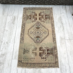 May include: A rectangular, vintage-style rug with a beige background and intricate geometric patterns in brown and teal. The rug features a central diamond shape and borders with repeating designs. The rug is placed on a light-colored wooden floor.