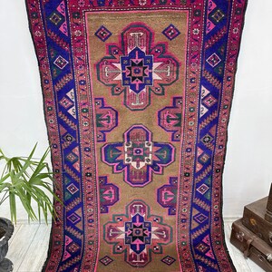 May include: A vintage Turkish rug with a brown background and a repeating pattern of pink, blue, and green geometric shapes. The rug has a fringe border.