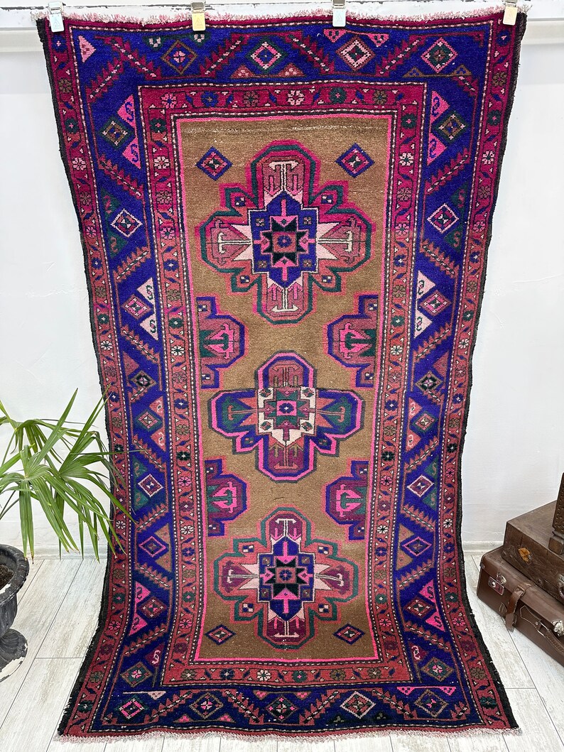 May include: A vintage Turkish rug with a brown background and a repeating pattern of pink, blue, and green geometric shapes. The rug has a fringe border.