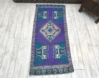 Puede incluir: Una alfombra rectangular tipo pasillero con un campo de color morado vibrante y un medallón central en forma de diamante en gris, blanco y verde azulado. Presenta motivos geométricos simétricos y un borde decorativo en gris y morado.