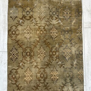 May include: A rectangular area rug featuring a muted olive green and brown color palette. The surface displays a repeating geometric pattern composed of interconnected circular motifs and stylized floral shapes arranged in a vertical grid layout.