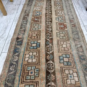 May include: A rectangular, vintage rug with a geometric pattern. The rug features vertical stripes in shades of brown, beige, and blue. The design includes repeating square and floral motifs. The rug appears to be made of woven fabric.