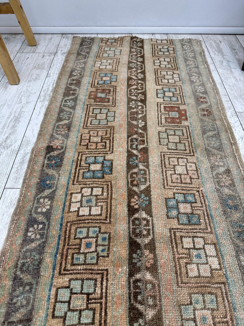 May include: A rectangular, vintage rug with a geometric pattern. The rug features vertical stripes in shades of brown, beige, and blue. The design includes repeating square and floral motifs. The rug appears to be made of woven fabric.