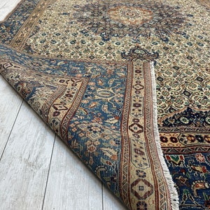 May include: A vintage Persian rug with a cream background and intricate blue, red, and brown floral patterns. The rug is folded in half, revealing the underside.