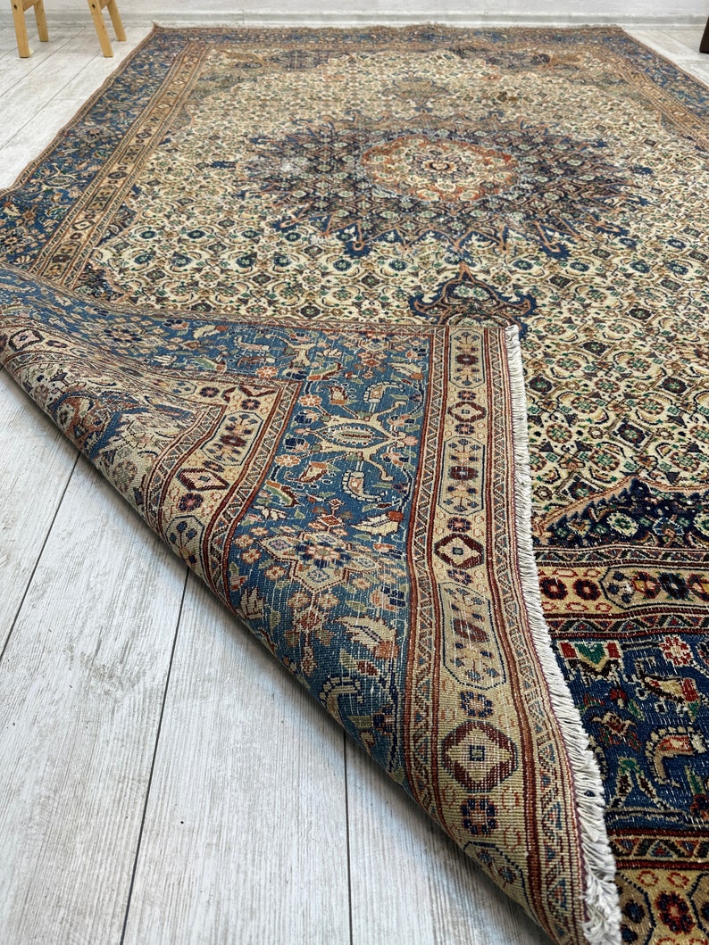 May include: A vintage Persian rug with a cream background and intricate blue, red, and brown floral patterns. The rug is folded in half, revealing the underside.