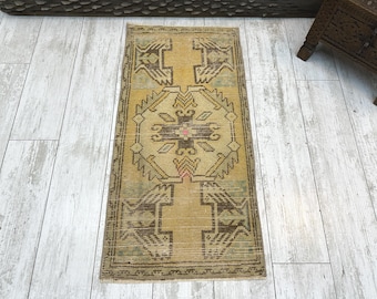 Puede incluir: Una alfombra tejida rectangular con fondo amarillo mostaza y un medallón geométrico central en marrón oscuro y verde apagado. El diseño incluye motivos angulares simétricos en los extremos con un pequeño acento de color rosa.