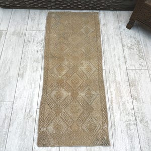 May include: A rectangular runner rug featuring a faded tan and beige colour palette with a repeating geometric pattern of diamonds and interconnected lines. The textile has a worn, textured appearance with visible weave details throughout the surface.