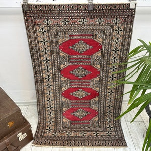 May include: A vintage rug with a geometric pattern in shades of red, black, and cream. The rug has a traditional design with intricate details.