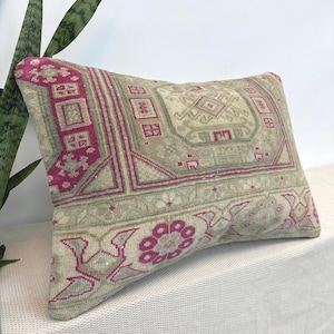 May include: A rectangular throw pillow featuring a woven textile pattern in shades of muted beige, light gray, and vibrant magenta. The design includes geometric borders, central medallion motifs, and floral-inspired shapes across the surface.