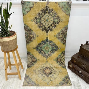 Puede incluir: Una alfombra vintage con fondo amarillo y intrincados patrones florales en tonos azules, verdes y marrones. La alfombra tiene un aspecto desgastado, lo que le da un toque único y antiguo.