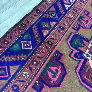 May include: A vintage rug with a brown background and a colorful geometric pattern in shades of pink, blue, green, and black. The rug has a fringed edge.