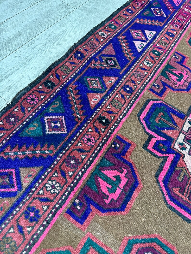 May include: A vintage rug with a brown background and a colorful geometric pattern in shades of pink, blue, green, and black. The rug has a fringed edge.