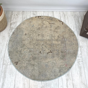 May include: A round area rug featuring a muted beige and gray color palette with a faded, intricate floral pattern. The rug has a distressed texture with visible dark brown accents scattered across the surface, creating a weathered appearance.