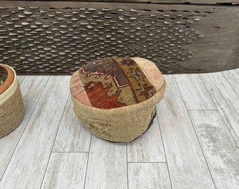 May include: A circular patchwork ottoman with a beige, patterned base and a top featuring a mix of colours including red, brown, and pink. The ottoman sits on a light wood floor.