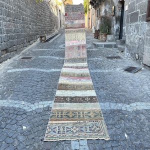 Puede incluir: Una alfombra vintage larga y rectangular con un diseño de patchwork. La alfombra presenta varios colores, incluyendo beige, rosa, marrón y azul, con patrones geométricos y florales. La alfombra se exhibe al aire libre en una calle adoquinada.