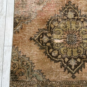 Puede incluir: Una alfombra vintage con un fondo marr&oacute;n descolorido y un gran dise&ntilde;o floral verde y negro intrincado en el centro. La alfombra tiene un aspecto desgastado y descolorido y un borde con flecos.