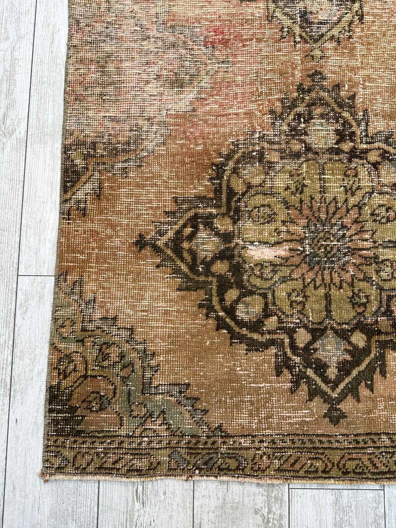 Puede incluir: Una alfombra vintage con un fondo marr&oacute;n descolorido y un gran dise&ntilde;o floral verde y negro intrincado en el centro. La alfombra tiene un aspecto desgastado y descolorido y un borde con flecos.
