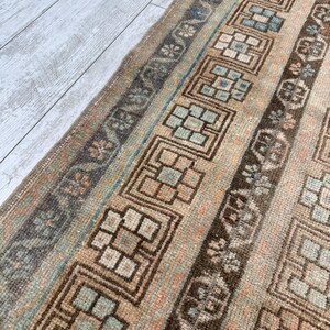 May include: A vintage rug with a repeating geometric pattern in shades of brown, beige, and blue. The rug features a central design with square and rectangular shapes, bordered by floral and linear elements. The rug's colors are muted, giving it a worn, antique appearance.