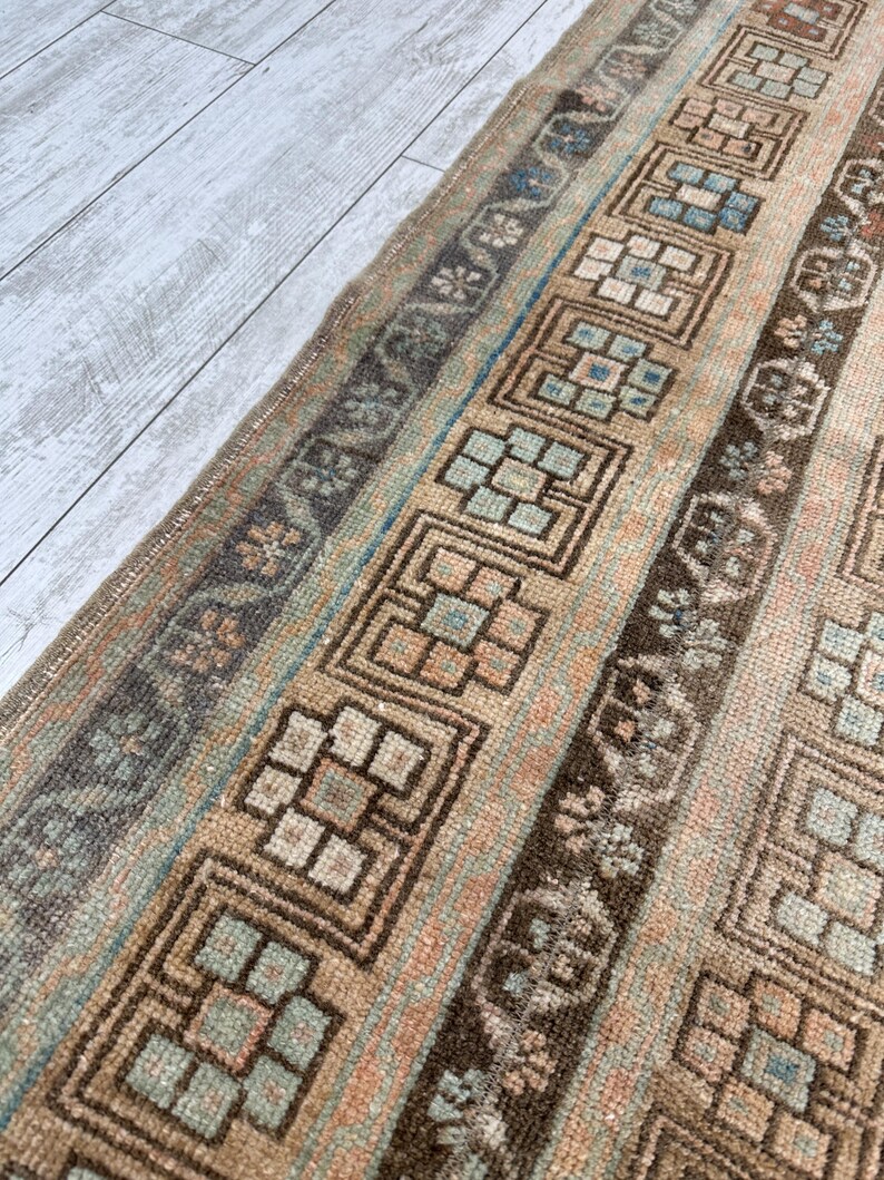 May include: A vintage rug with a repeating geometric pattern in shades of brown, beige, and blue. The rug features a central design with square and rectangular shapes, bordered by floral and linear elements. The rug's colors are muted, giving it a worn, antique appearance.