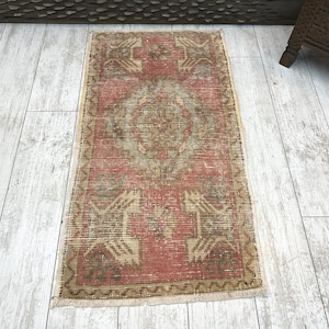 Puede incluir: Una alfombra rectangular vintage con un aspecto descolorido y desgastado. La alfombra presenta un dise&ntilde;o de medall&oacute;n central en tonos beige y verde sobre un fondo rojo. La alfombra tiene un borde y flecos beige.