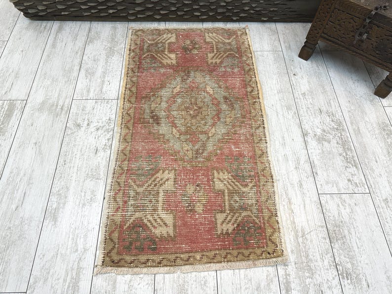 Puede incluir: Una alfombra rectangular vintage con un aspecto descolorido y desgastado. La alfombra presenta un dise&ntilde;o de medall&oacute;n central en tonos beige y verde sobre un fondo rojo. La alfombra tiene un borde y flecos beige.