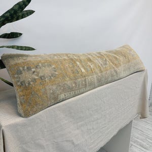May include: A long rectangular lumbar pillow featuring a faded vintage-style woven pattern in shades of mustard yellow, muted gray, and cream. The textile displays intricate geometric and floral motifs across its surface with a textured finish.