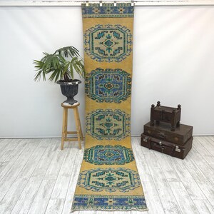 May include: A long, narrow runner rug featuring a vibrant mustard yellow background with five repeating geometric medallions in shades of teal, navy blue, and cream. The rug has a decorative blue border at both ends with intricate patterns.