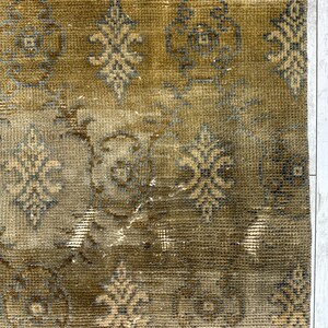 May include: A rectangular area rug featuring a muted olive green and brown base color with a repeating pattern of dark gray and light tan floral motifs. The rug has a textured, woven surface with a frayed edge along the top border.