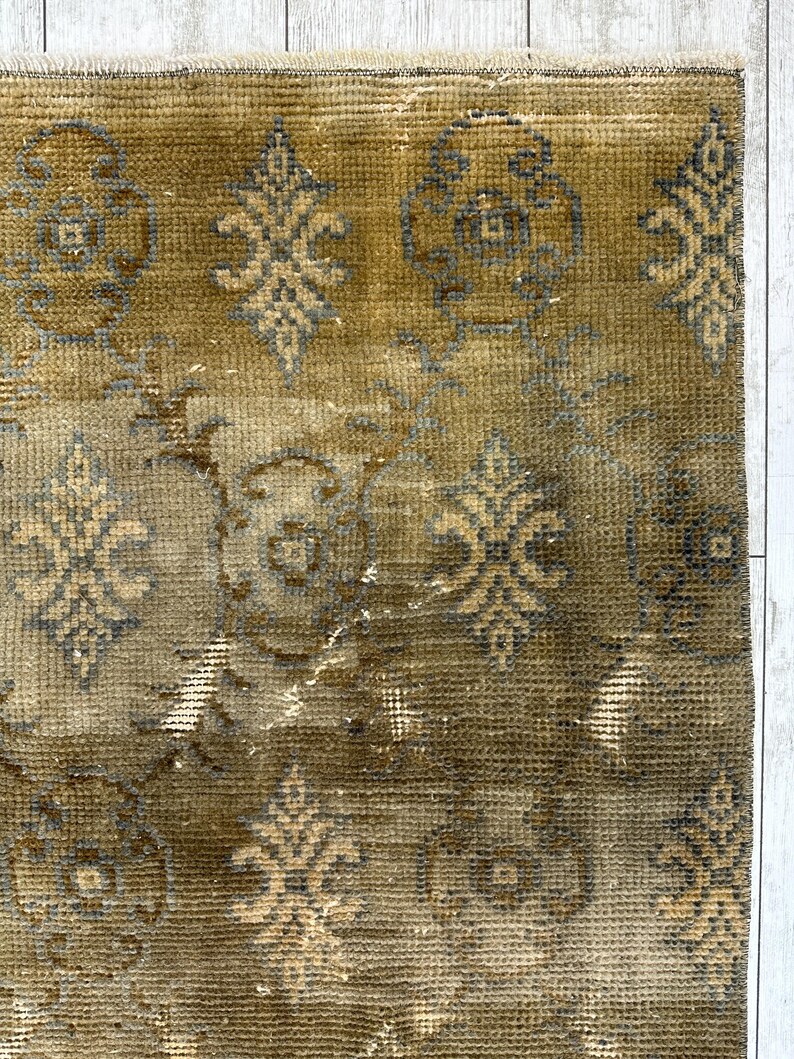 May include: A rectangular area rug featuring a muted olive green and brown base color with a repeating pattern of dark gray and light tan floral motifs. The rug has a textured, woven surface with a frayed edge along the top border.