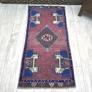 May include: A rectangular, vintage rug with a faded red background and intricate geometric patterns. The rug features blue, white, and brown accents, with a central diamond-shaped design. The border is a dark blue with repeating patterns.