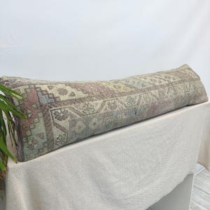 May include: A long, rectangular lumbar pillow featuring a woven textile cover with a faded, intricate geometric pattern in muted shades of dusty rose, sage green, light blue, and cream. The fabric displays a weathered, textured appearance.