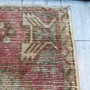 Puede incluir: Primer plano de una secci&oacute;n de una alfombra vintage con un aspecto descolorido y desgastado. La alfombra presenta un patr&oacute;n geom&eacute;trico repetitivo en tonos de rojo, beige y verde. Los bordes est&aacute;n deshilachados, lo que se suma a su est&eacute;tica envejecida. La alfombra est&aacute; colocada sobre una superficie de madera clara.