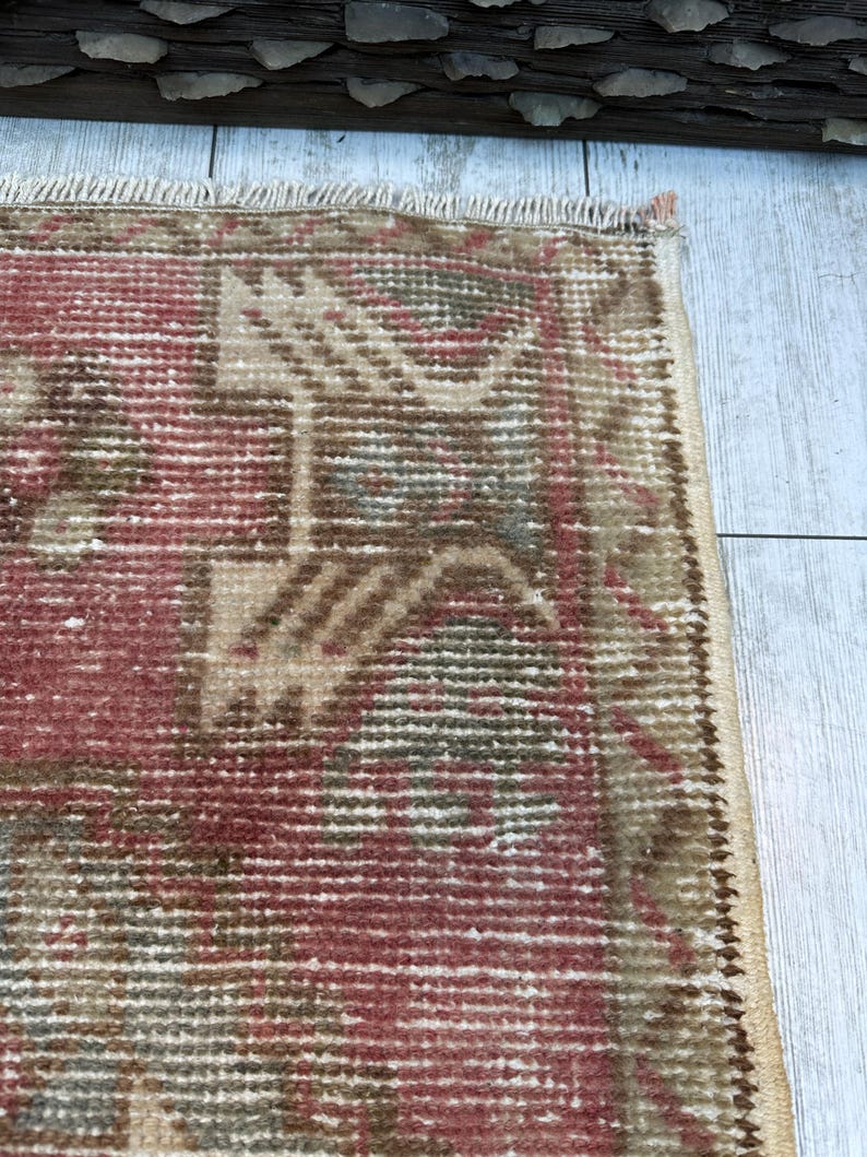 Puede incluir: Primer plano de una secci&oacute;n de una alfombra vintage con un aspecto descolorido y desgastado. La alfombra presenta un patr&oacute;n geom&eacute;trico repetitivo en tonos de rojo, beige y verde. Los bordes est&aacute;n deshilachados, lo que se suma a su est&eacute;tica envejecida. La alfombra est&aacute; colocada sobre una superficie de madera clara.