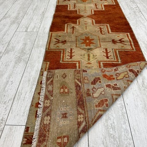 May include: A vintage Turkish runner rug with a geometric pattern in shades of red, brown, and beige. The rug has a fringed edge and is folded in half.