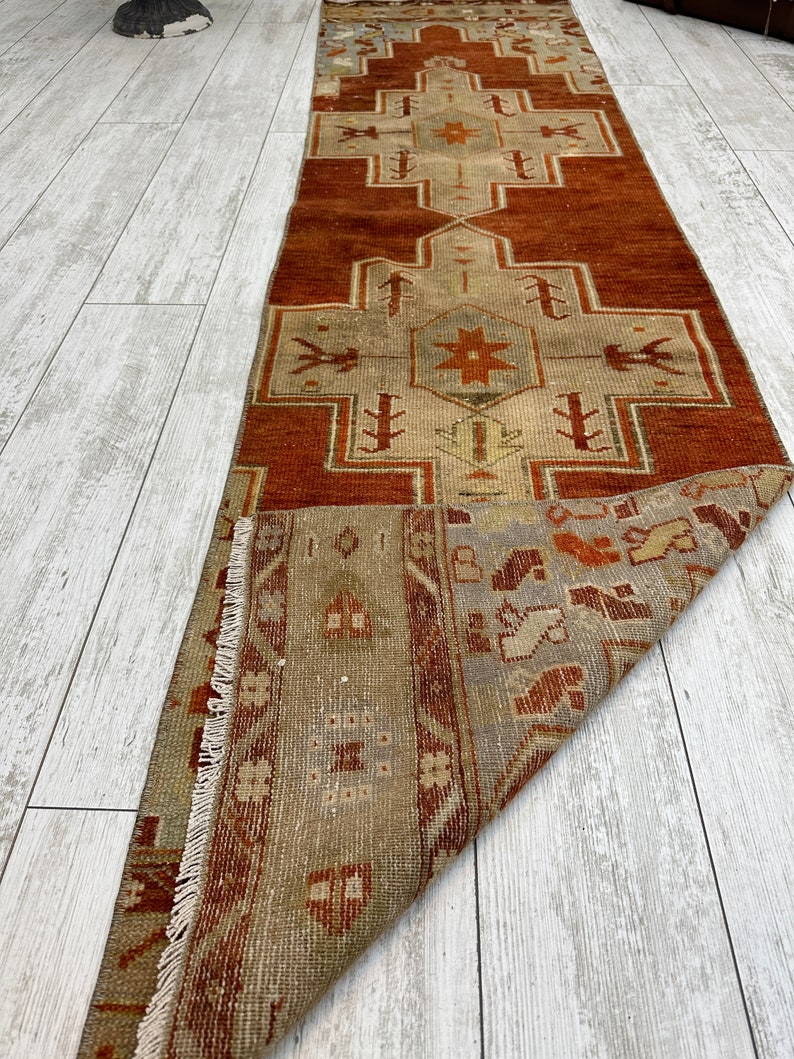 May include: A vintage Turkish runner rug with a geometric pattern in shades of red, brown, and beige. The rug has a fringed edge and is folded in half.