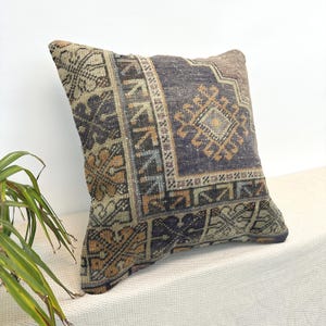 May include: A square throw pillow featuring a woven textile cover with a dark purple, tan, and muted orange geometric pattern. The design includes intricate borders and a central diamond motif, showcasing a weathered, textured appearance.