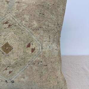 May include: A square throw pillow featuring a textured, woven surface in muted beige and cream tones. The fabric displays a faded, intricate geometric pattern with small accents of soft brown, dusty pink, and pale green throughout the weave.