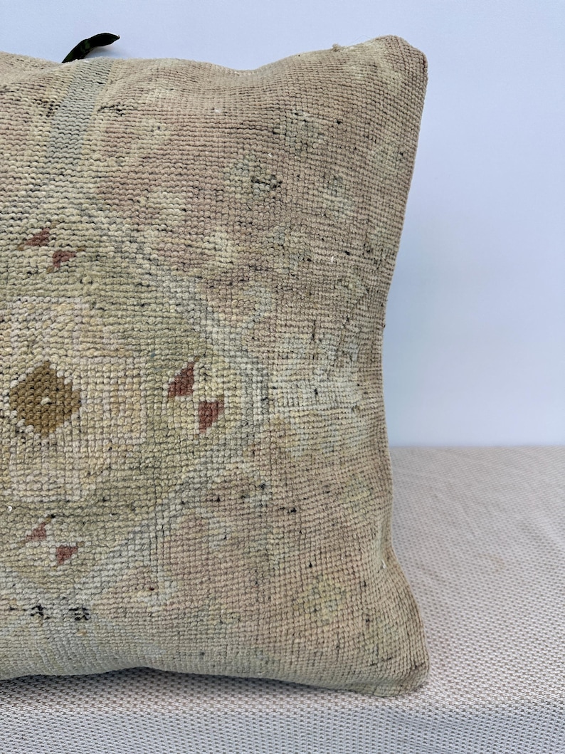 May include: A square throw pillow featuring a textured, woven surface in muted beige and cream tones. The fabric displays a faded, intricate geometric pattern with small accents of soft brown, dusty pink, and pale green throughout the weave.