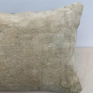 May include: A rectangular throw pillow featuring a woven, textured surface in a muted beige and tan colour palette. The fabric displays a dense, grid-like pattern with subtle variations in tone and slight signs of wear, creating a weathered appearance.
