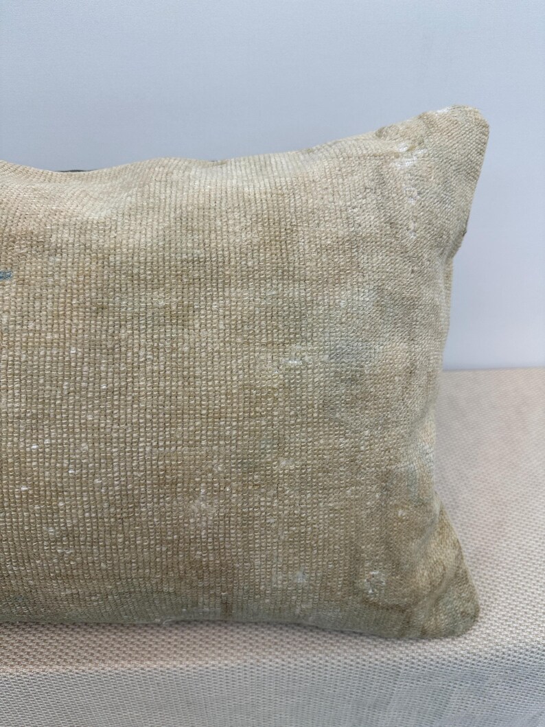 May include: A rectangular throw pillow featuring a woven, textured surface in a muted beige and tan colour palette. The fabric displays a dense, grid-like pattern with subtle variations in tone and slight signs of wear, creating a weathered appearance.