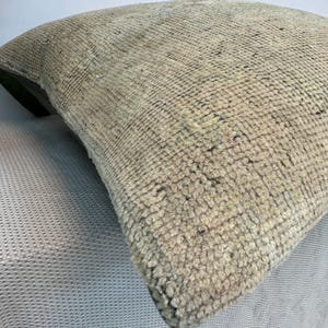 May include: A square throw pillow featuring a thick, hand-knotted wool pile in a neutral beige and tan color palette. The textured surface displays a dense, looped weave with subtle variations in tone, creating a tactile and uniform appearance.
