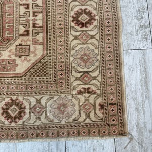May include: Close-up of the bottom edge of a beige rug with a brown and pink geometric pattern. The rug has a fringe border.